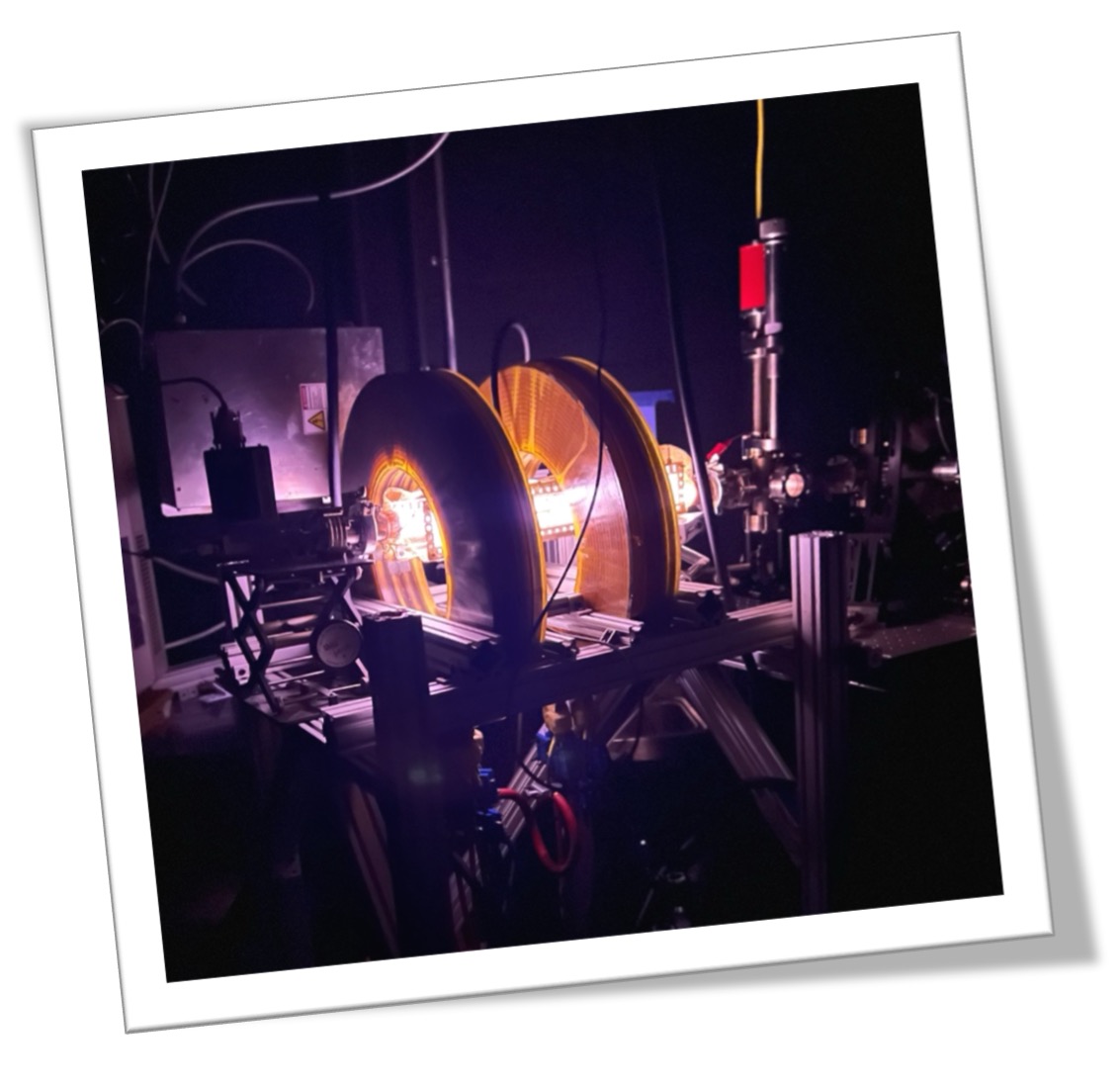 A close-up view of a scientific apparatus featuring two circular components illuminated in purple and yellow light, set against a dark background, with various connected instruments and tubing.